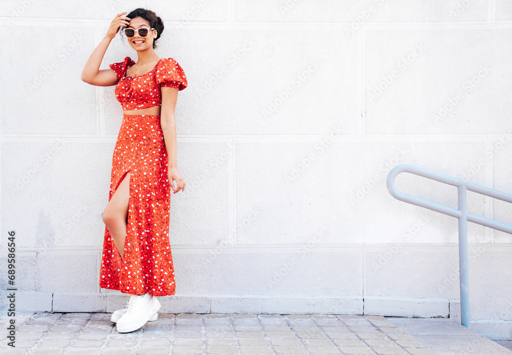 Young beautiful smiling hipster woman in trendy summer red dress. Carefree woman posing in the street near wall. Positive model outdoors in at sunny day. Cheerful and happy. In sunglasses Young beautiful smiling hipster woman in trendy summer red dress. Carefree woman posing in the street near wall. Positive model outdoors in at sunny day. Cheerful and happy. In sunglasses