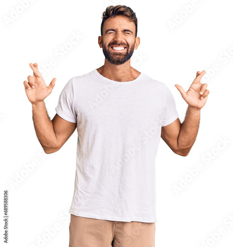 Young hispanic man wearing casual white tshirt gesturing finger crossed smiling with hope and eyes closed. luck and superstitious concept.