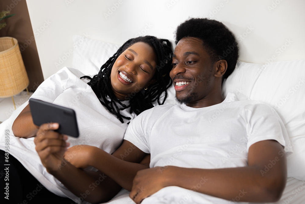 Fototapeta premium Delighted happy american black family sit on bed with cell phone smiling look at smartphoone screen making video call with friends, using new mobile application, watch videos.