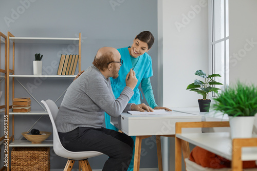 Friendly nurse taking care of a senior man in a retirement home. Happy, smiling young woman helping an old man who is sitting at his desk, taking notes in his diary or writing a letter to his family