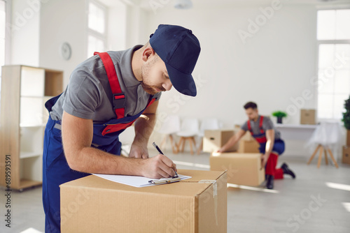 Worker from professional man and van delivery company writes something on paper while removing stuff on house moving day. Loader in uniform workwear and baseball cap fills out invoice on cardboard box