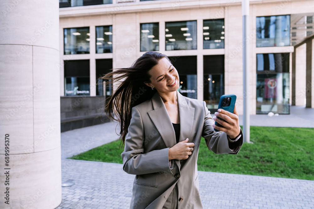Cheerful young woman in business suit makes video call by phone walking by street toothy smiles happy goes home after work. Female employee happy outside. Cheerful young woman in business suit makes video call by phone walking by street toothy smiles happy goes home after work. Female employee happy outside.
