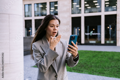 Shocked young woman in suit holds phone looks at screen in surprise, frustrated by bad news standing outside. Unhappy businesswoman received information about financial crisis in company.