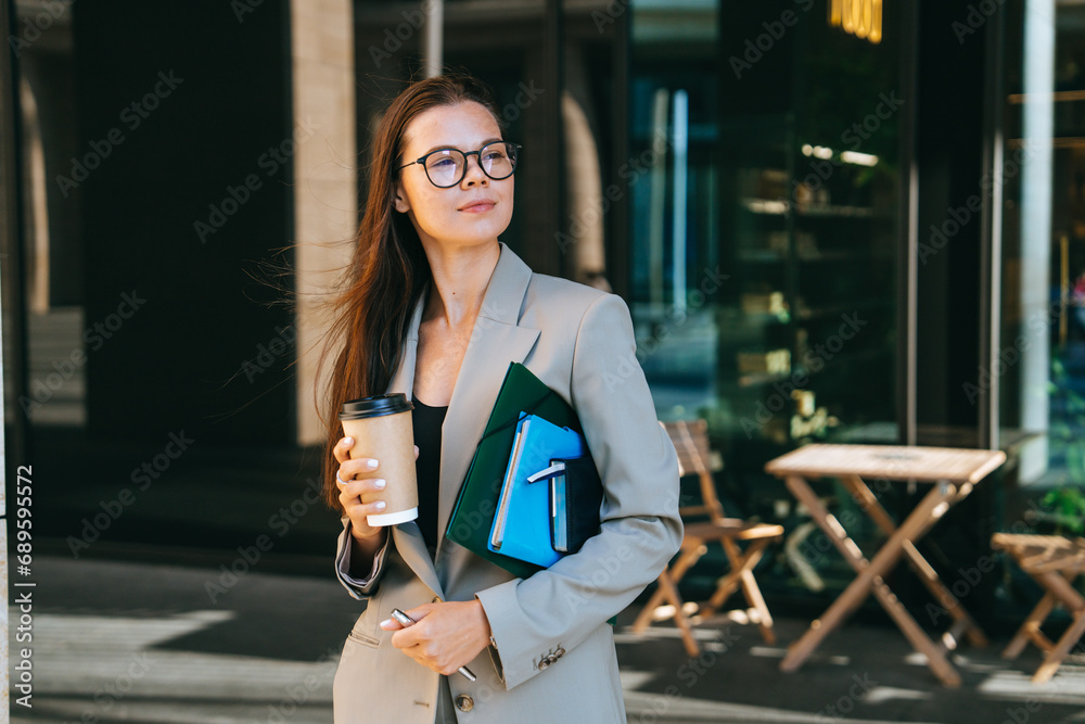 Thoughtful brunette businesswoman in suit holds coffee cup, notebooks, documents relaxing, having break need reboot. American student girl in glasses goes to university.