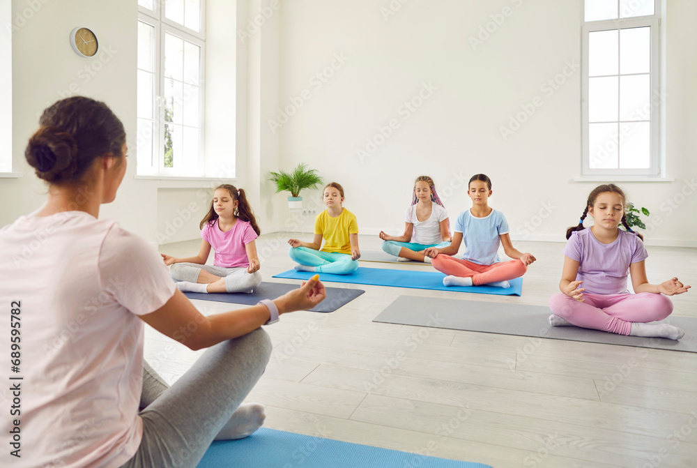 Group of kids having a yoga lesson. Children doing relaxing exercises. Little girls in sportswear sitting on the floor on mats in the lotus pose and meditating together with their teacher Group of kids having a yoga lesson. Children doing relaxing exercises. Little girls in sportswear sitting on the floor on mats in the lotus pose and meditating together with their teacher