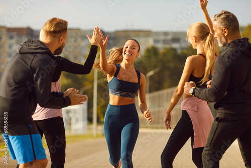 Fototapeta Naklejka Na Ścianę i Meble -  Happy smiling sporty young friends giving high five to a woman rejoicing the success in sport training after jogging exercises outdoors. People standing in the park after workout in nature.