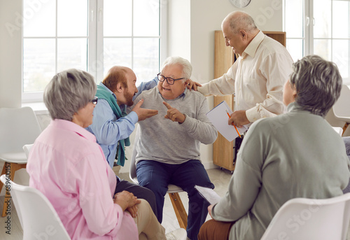 Two old men argue, exchange negative opinions, offend, insult each other while sitting with other senior people at talking therapy session devoted to constructive communication and conflict resolution photo