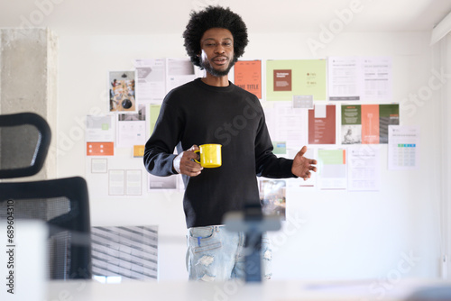 Afro man with casual style in his office talking and gesturing during a video call while holding a cup of coffee. In the background is the moodboard with projects in progress of the business. In the f photo