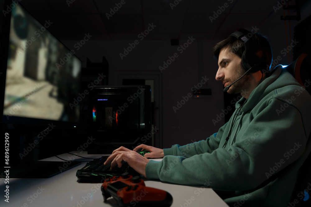 Side view of concentrated male eSport gamer in headphones with microphone sitting at white table and playing video game on desktop in dark room Side view of concentrated male eSport gamer in headphones with microphone sitting at white table and playing video game on desktop in dark room