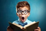 Surprised little boy in glasses with backpack reading book on blue background. Back to school concept