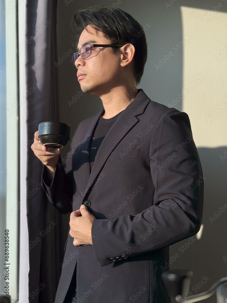 Naklejka premium Vertical portrait of an Asian man with short, cool black hair, wearing glasses, wearing a black suit and businessman, drinking coffee with a shadow of a window in the background.