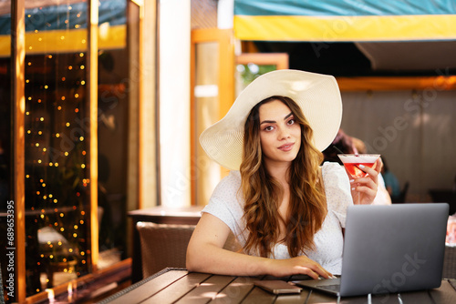 Female freelancer in summer hat browsing laptop at table while working on remote project in outdoor cafe, having break and drinking cocktail