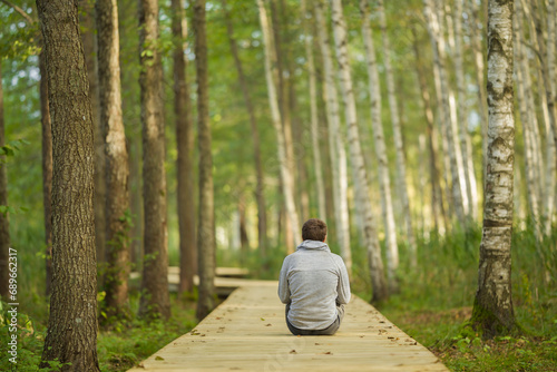 Young adult man sitting on wooden trail at birch tree forest. Looking far away. Spending time alone in beautiful autumn day. Back view. Thinking about life. Peaceful atmosphere in nature.