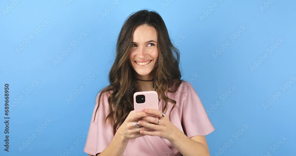 Smiling beautiful young woman isolated on blue background. Girl in pink dress flirting while chatting on mobile phone, dreaming. Woman think about answer, bite lip, look at side. Smiling beautiful young woman isolated on blue background. Girl in pink dress flirting while chatting on mobile phone, dreaming. Woman think about answer, bite lip, look at side.