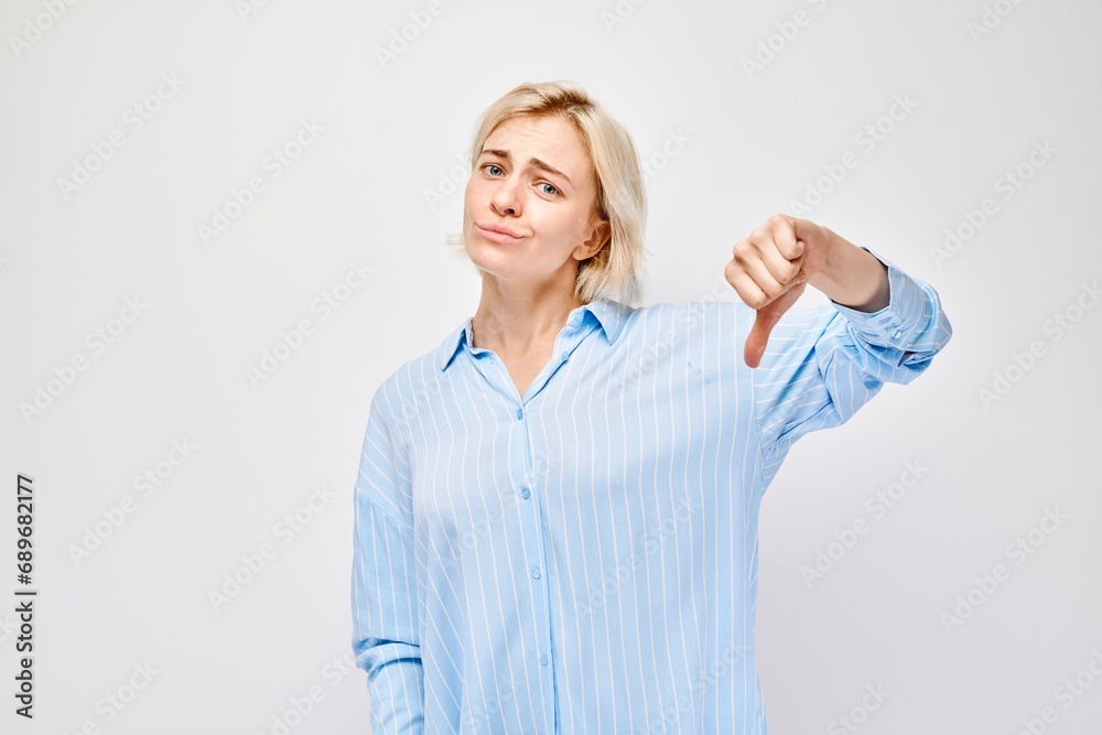 Obraz premium Portrait of blond girl in casual shirt showing thumbs down isolated on white studio background. Dislike, wrong choice, negative evaluation concept.
