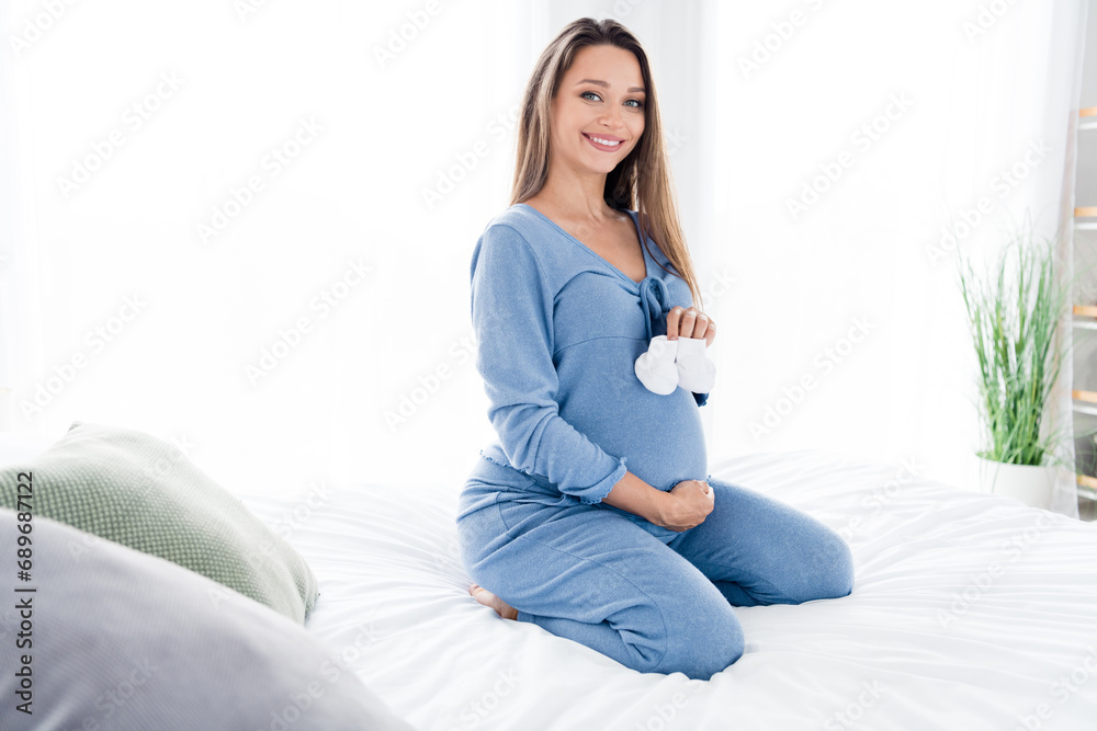 Photo of adorable cute mom preparing for childbirth showing holding tiny small socks sitting in bed white light room indoors Photo of adorable cute mom preparing for childbirth showing holding tiny small socks sitting in bed white light room indoors
