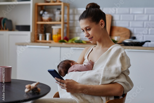 Side view at happy Caucasian mother sitting in kitchen using smartphone while holding napping baby in blanket, copy space photo