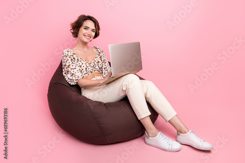 Full length photo of optimistic cute woman wear print top sit on bean bag texting in chat on laptop isolated on pink color background