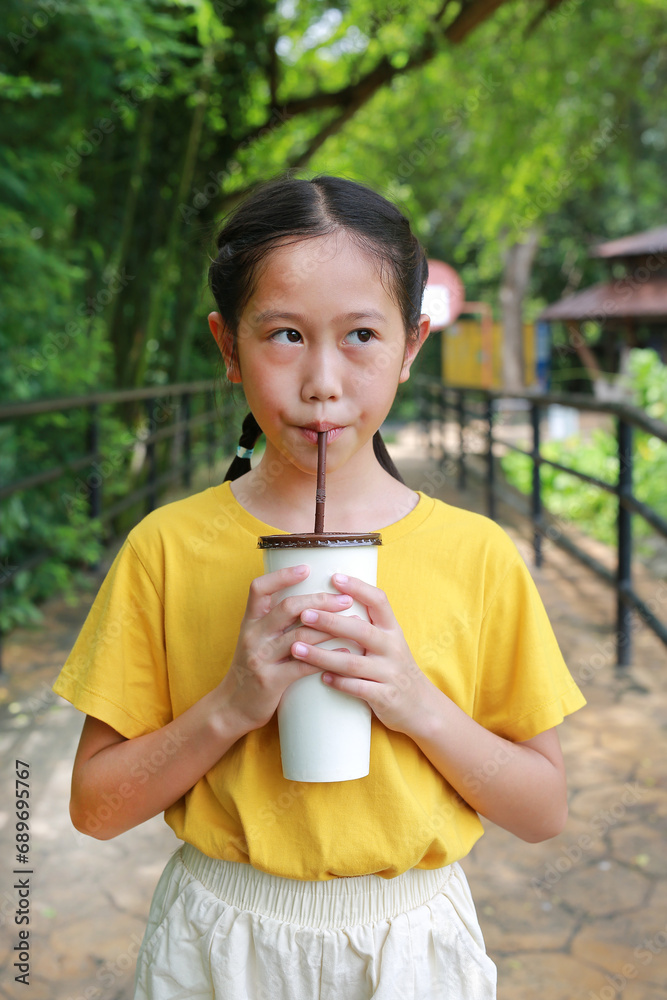 Portrait of Asian young girl child holding paper cup and drinking cold water by straw with looking beside while standing in public garden. Portrait of Asian young girl child holding paper cup and drinking cold water by straw with looking beside while standing in public garden.