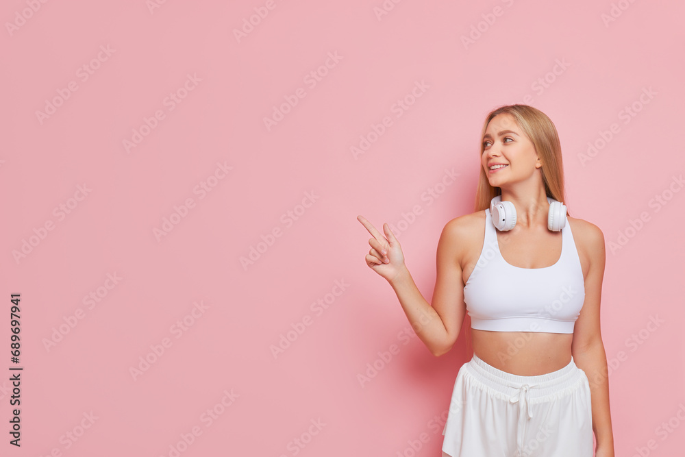 Smiling girl with blond hair in white short top poses on pink backdrop with index finger up, nice day concept, copy space Smiling girl with blond hair in white short top poses on pink backdrop with index finger up, nice day concept, copy space