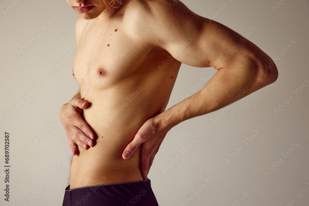 Cropped image of muscular shirtless male body against grey studio background. Young man holding hands on lower back. Pains, injury. Concept of men's beauty, health, body care, sportive lifestyle