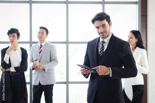 Portrait of young confident and smart businessman in black suit using digital tablet while confident standing at modern office with colleague team as blurred background. Happy officer at workplace.