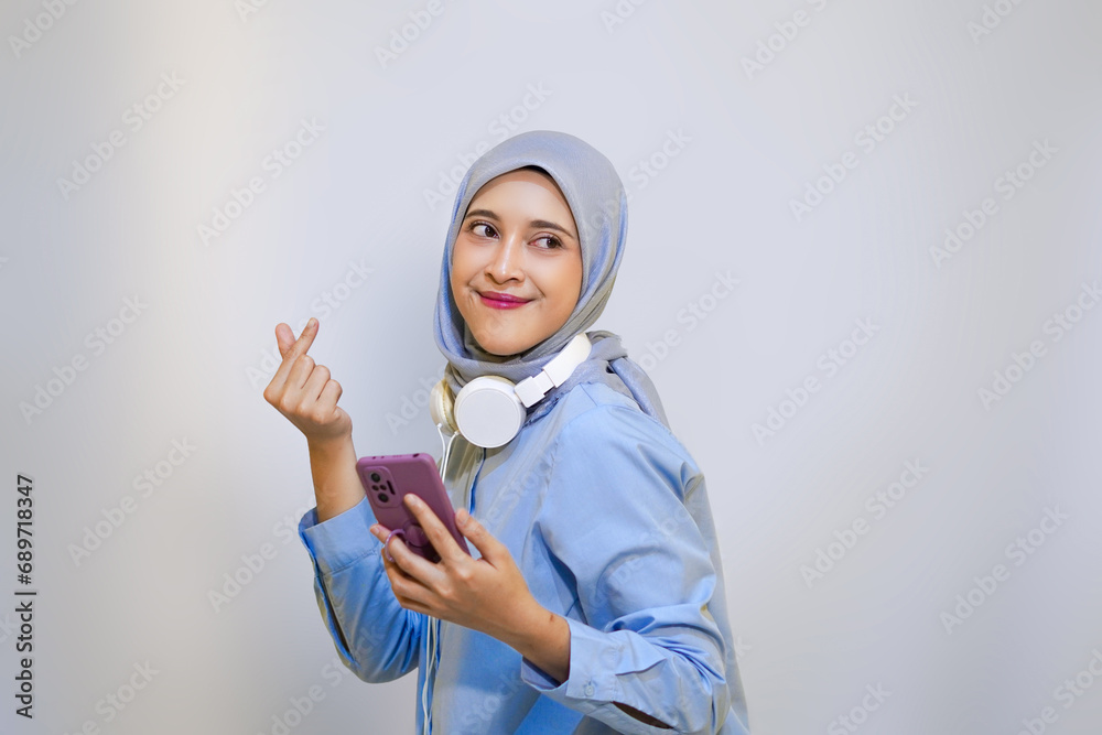Cheerful muslim woman with love sign finger loves the music on her mobile phone. wired headphone and listening music concept Cheerful muslim woman with love sign finger loves the music on her mobile phone. wired headphone and listening music concept
