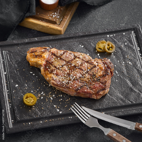 Juicy grilled striploin steak seasoned with coarse salt and black pepper, accompanied by jalapeno slices on a black slate surface