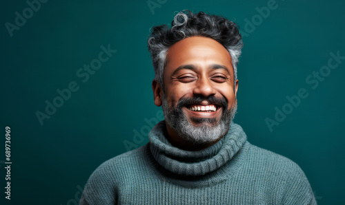 Portrait of a joyful middle-aged South Asian man with salt and pepper hair, smiling in a grey sweater against a teal background