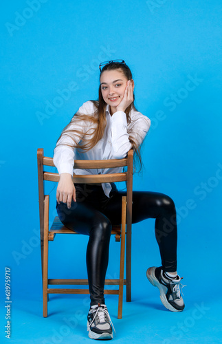 Front view of smiling, young caucasian woman with long fair hair and sensual gaze resting head on hand, while sitting on chair backwards and looking at camera, isolated on blue. Concept of posing.
