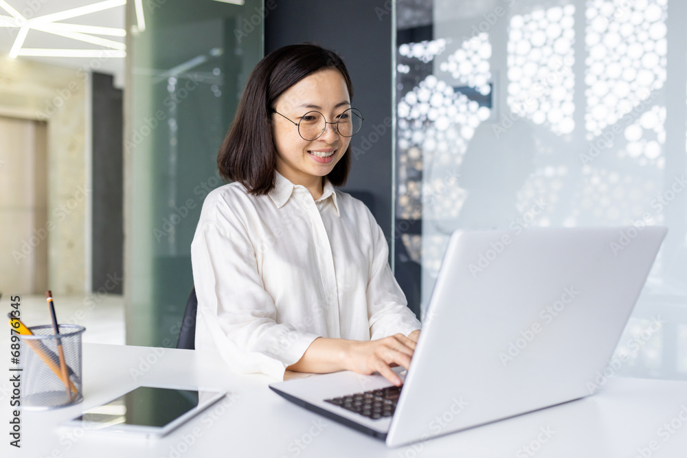 Successful Asian business woman at workplace inside office, female employee smiling contentedly using laptop at work, woman in light clothes satisfied with achievement results, typing on keyboard. Successful Asian business woman at workplace inside office, female employee smiling contentedly using laptop at work, woman in light clothes satisfied with achievement results, typing on keyboard.