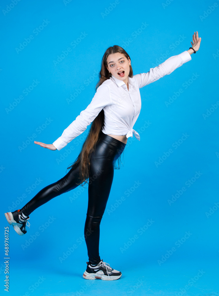 Obraz premium Excited young woman in white shirt raising arm and leg, while posing indoors. Side view of joyful girl outstretching hands, while turning to camera, isolated on blue background. Concept of happiness.