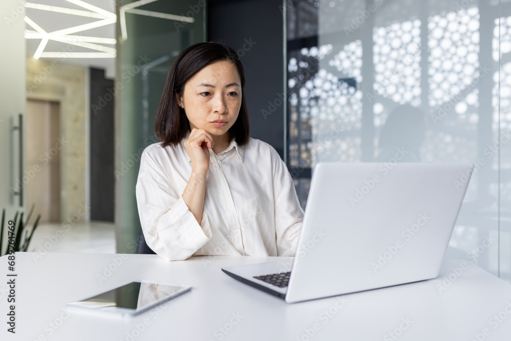 Serious thinking asian woman inside office at workplace, business woman with laptop reading data from monitor, hand on chin, brainstorming strategy plan and proofreading preparing financial report. Serious thinking asian woman inside office at workplace, business woman with laptop reading data from monitor, hand on chin, brainstorming strategy plan and proofreading preparing financial report.