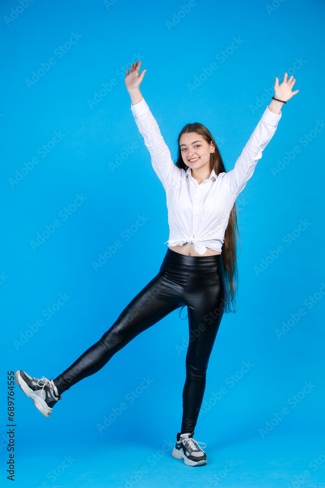 Obraz premium Cheerful young lady in skinny leggings raising arms and leg, while posing in studio. Front view of happy girl keeping hands up, while looking at camera, isolated on blue background. Concept of fun.