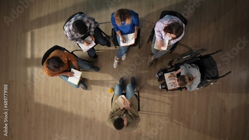 In the shot above, a group of people and their mentor are sitting on a chair. They have come for therapy. They are sitting in a circle writing something in a notebook with a disabled person photo