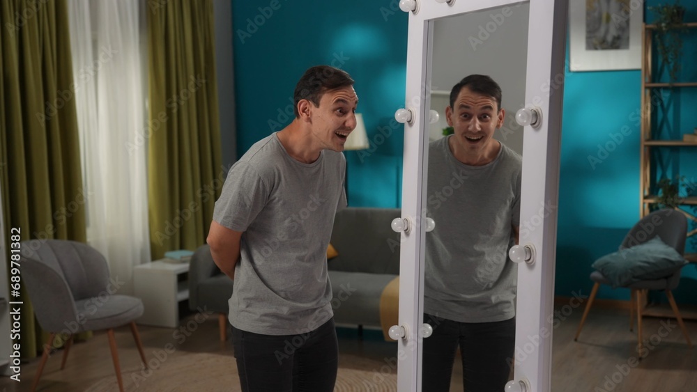 A man with bipolar disorder at a mirror in the living room. The man is writhing, laughing and mocking his reflection. Dark side, bipolar disorder, dissociative disorder. A man with bipolar disorder at a mirror in the living room. The man is writhing, laughing and mocking his reflection. Dark side, bipolar disorder, dissociative disorder.