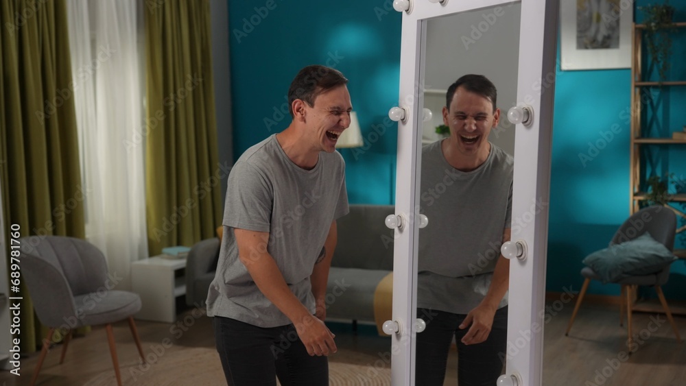 A man with bipolar disorder at a mirror in the living room. The man is writhing, laughing and mocking his reflection. Dark side, bipolar disorder, dissociative disorder. A man with bipolar disorder at a mirror in the living room. The man is writhing, laughing and mocking his reflection. Dark side, bipolar disorder, dissociative disorder.