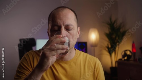 Middle aged man takes pill with glass of water in hand. Stressed mature man drinking sedated antidepressant meds. Man feels depressed, taking drugs. Medicines at home, 4k video footage, medium shot photo