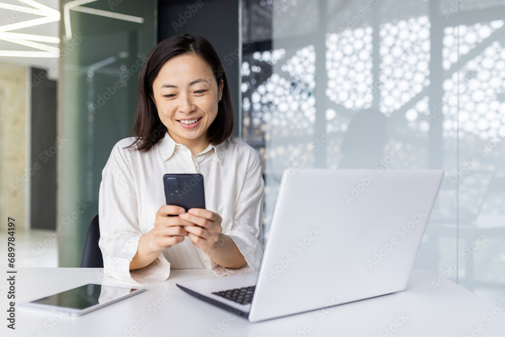 Young beautiful woman at workplace holds phone in her hands, Asian business woman reads online e-mail, female worker with a laptop inside the office browses the Internet pages, smiles contentedly. Young beautiful woman at workplace holds phone in her hands, Asian business woman reads online e-mail, female worker with a laptop inside the office browses the Internet pages, smiles contentedly.