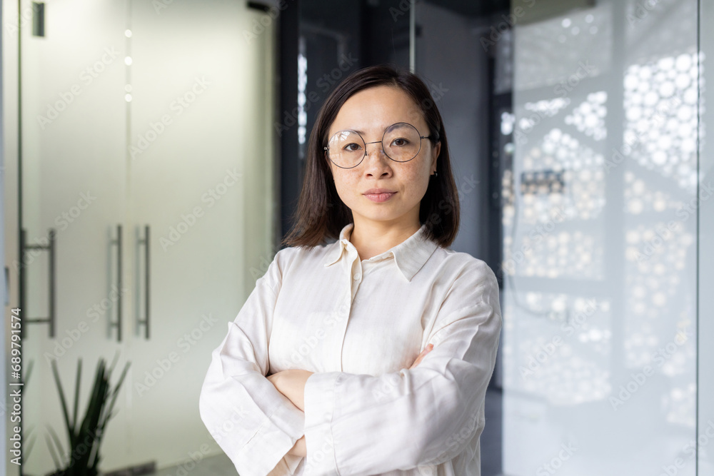 Portrait of mature experienced asian businesswoman boss, female worker looking at camera seriously, woman in glasses with crossed arms inside office building confident of successful achievement. Portrait of mature experienced asian businesswoman boss, female worker looking at camera seriously, woman in glasses with crossed arms inside office building confident of successful achievement.