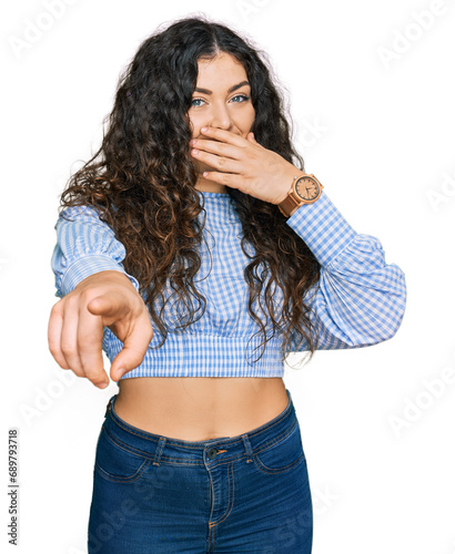 Young hispanic girl wearing casual clothes laughing at you, pointing finger to the camera with hand over mouth, shame expression