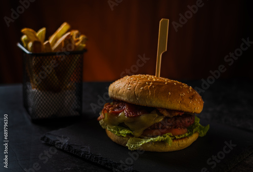 Fotografía de una hamburguesa de vacuno con queso, bacon, lechuga y tomate, con patatas fritas y fondo naranja, sobre superficie de pizarra.