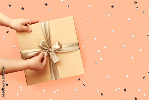 Children hands are holding a box with a gift. Cute festive peach fuzz trendy color background with sparkles. Conceptual backdrop for christmas, new year or birthday.