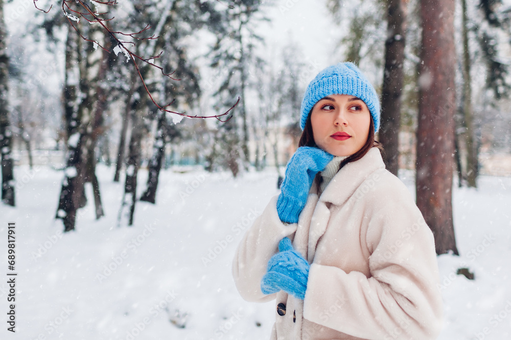 Close up portrait of young woman walking under snowfall in winter park wearing white fur coat blue hat mittens