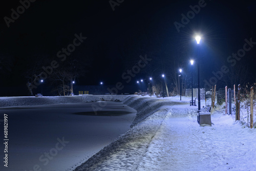 Staw nocą, w zimowej scenerii. Powierzchnnia stawu skuta lodem, brzeg pokryty warstwą białego śniegu. Wokół stawu znajduje się ścieżka przy której stoją ławki i latarnie. 