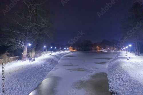 Staw nocą, w zimowej scenerii. Powierzchnnia stawu skuta lodem, brzeg pokryty warstwą białego śniegu. Wokół stawu znajduje się ścieżka przy której stoją ławki i latarnie. Zdjęcie z drona.
