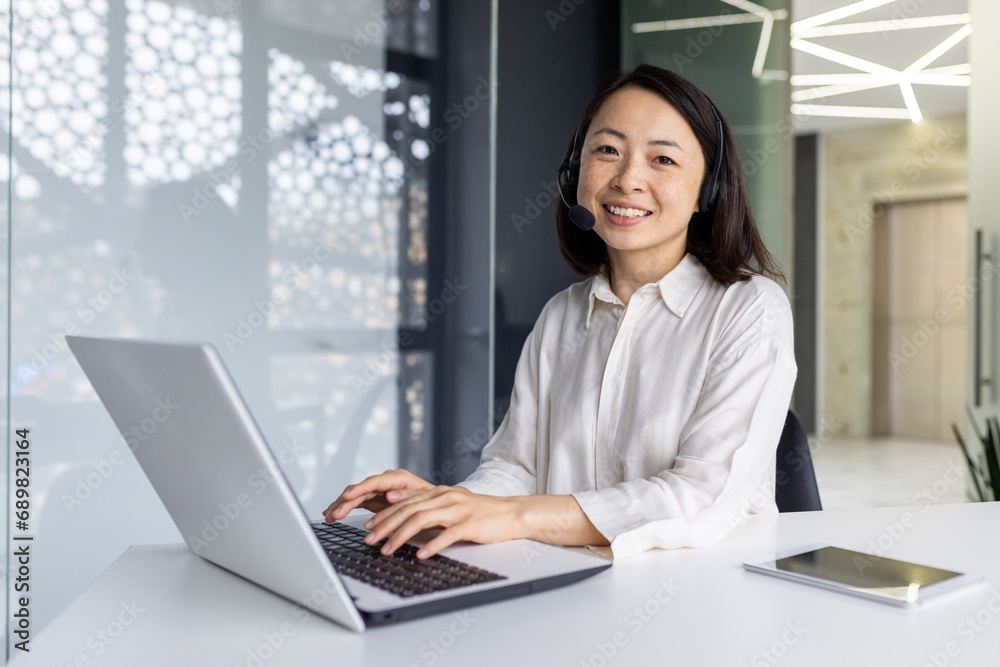 Portrait of online support worker, asian woman with headset phone smiling and looking at camera, businesswoman working in laptop at workplace inside office, call center call center support service. Portrait of online support worker, asian woman with headset phone smiling and looking at camera, businesswoman working in laptop at workplace inside office, call center call center support service.