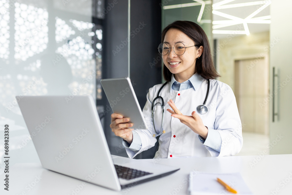 Young Asian female doctor working inside medical office consulting patients remotely, smiling with satisfaction at client, using tablet computer for video call, sitting at desk with laptop. Young Asian female doctor working inside medical office consulting patients remotely, smiling with satisfaction at client, using tablet computer for video call, sitting at desk with laptop.