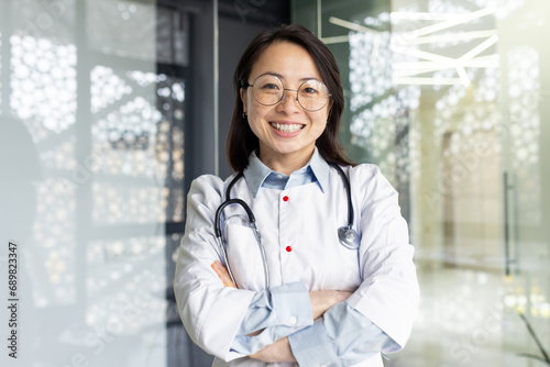 Portrait of a young beautiful and successful Asian woman inside a clinic office, a doctor smiling and looking at the camera with crossed arms and a white medical coat with a stethoscope.
