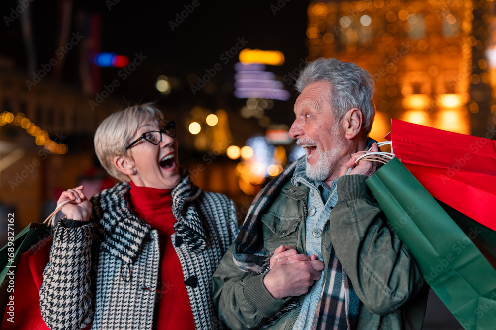 Excited senior couple looking at each other doing Christmas shopping walking with shopping bags. Two elderly people in good spirit after buying presents.
